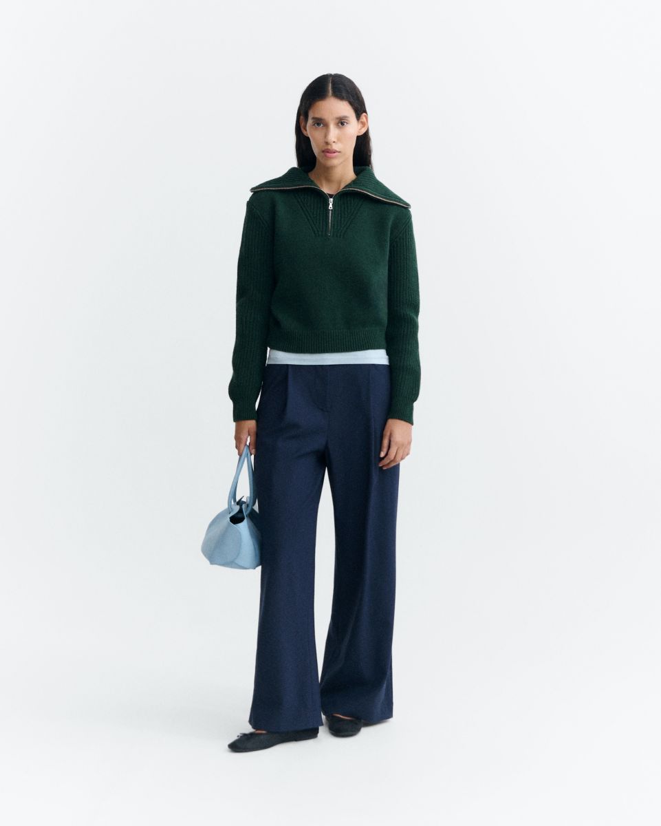 Dunkelgrüner Pullover "Tanit" von Thinking Mu aus Merinowolle mit hohem Kragen, silbernem Reißverschluss und Label am Ausschnitt vor hellem Hintergrund.