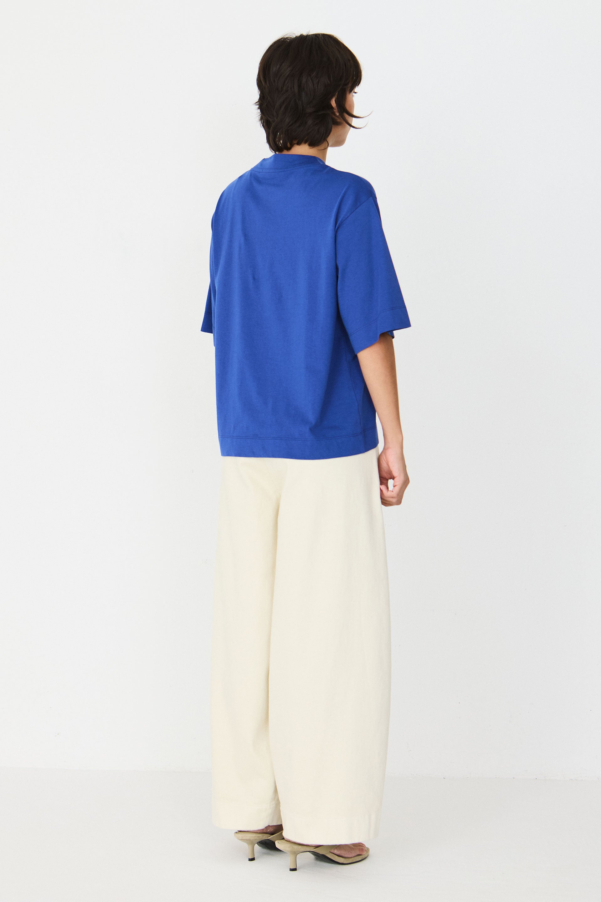 Model trägt das blaue Zuntz-11-Gots T-Shirt der Marke SKFK mit Stehkragen und kurzen Ärmeln zu hellen Hosen.
