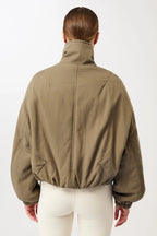 Eine Person trägt eine olivgrüne, sportive Blouson-Jacke mit hohem Stehkragen und Reißverschluss vor hellem Hintergrund