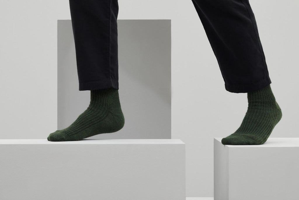 Ein Paar gestrickte Socken aus Wolle in meliertem Waldgrün vor weißem Hintergrund.