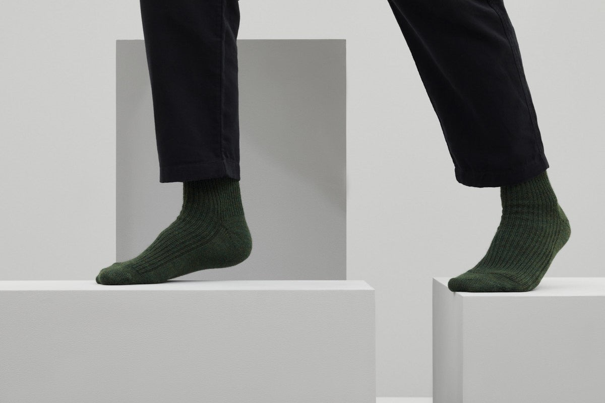 Ein Paar gestrickte Socken aus Wolle in meliertem Waldgrün vor weißem Hintergrund.