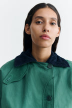 Grüne Jacke "Norma" von Thinking Mu mit dunklem Cord-Kragen, Knöpfen und herabhängenden Bändern am Saum vor hellem Hintergrund.