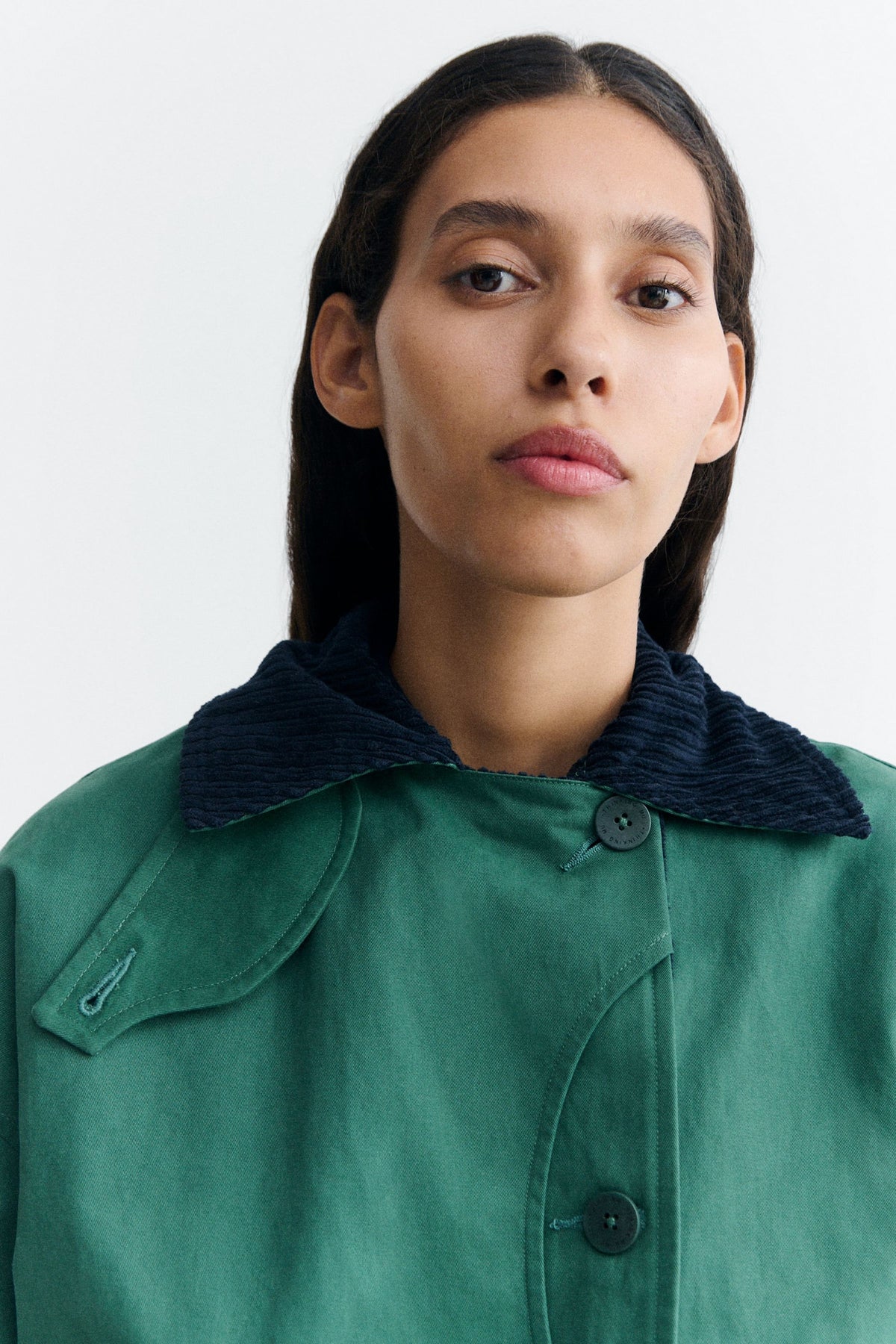 Grüne Jacke "Norma" von Thinking Mu mit dunklem Cord-Kragen, Knöpfen und herabhängenden Bändern am Saum vor hellem Hintergrund.