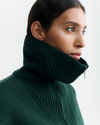Dunkelgrüner Pullover "Tanit" von Thinking Mu aus Merinowolle mit hohem Kragen, silbernem Reißverschluss und Label am Ausschnitt vor hellem Hintergrund.