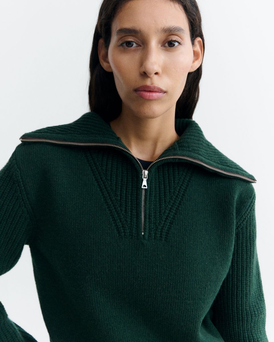 Dunkelgrüner Pullover "Tanit" von Thinking Mu aus Merinowolle mit hohem Kragen, silbernem Reißverschluss und Label am Ausschnitt vor hellem Hintergrund.