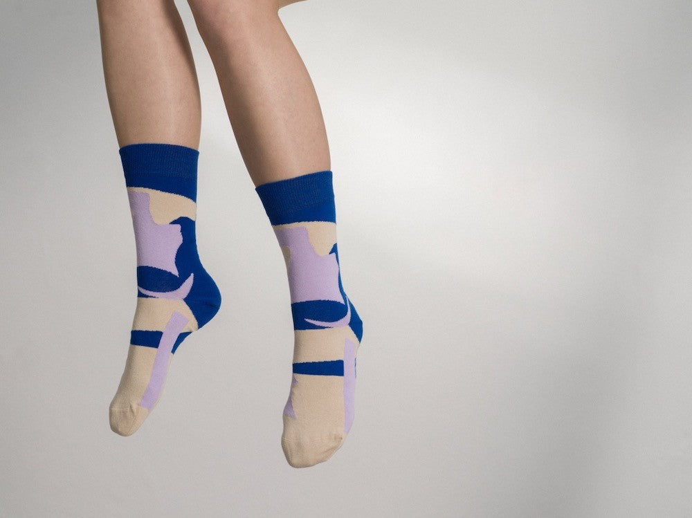 Ein Paar blaue Socken mit beigen und violetten, abstrakten Farb- und Blockmustern sowie beiger Fersen- und Zehenpartie vor hellem Hintergrund. koblenz baumwolle