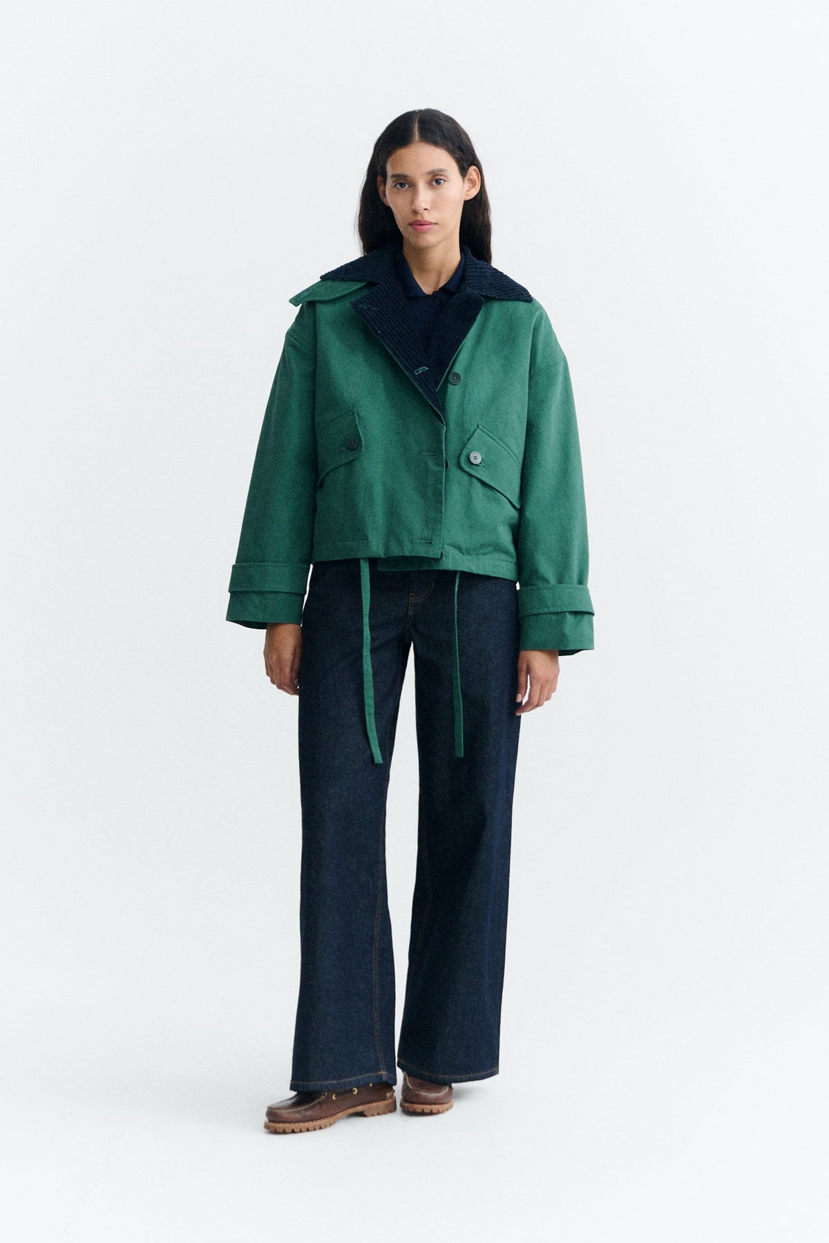 Grüne Jacke "Norma" von Thinking Mu mit dunklem Cord-Kragen, Knöpfen und herabhängenden Bändern am Saum vor hellem Hintergrund.