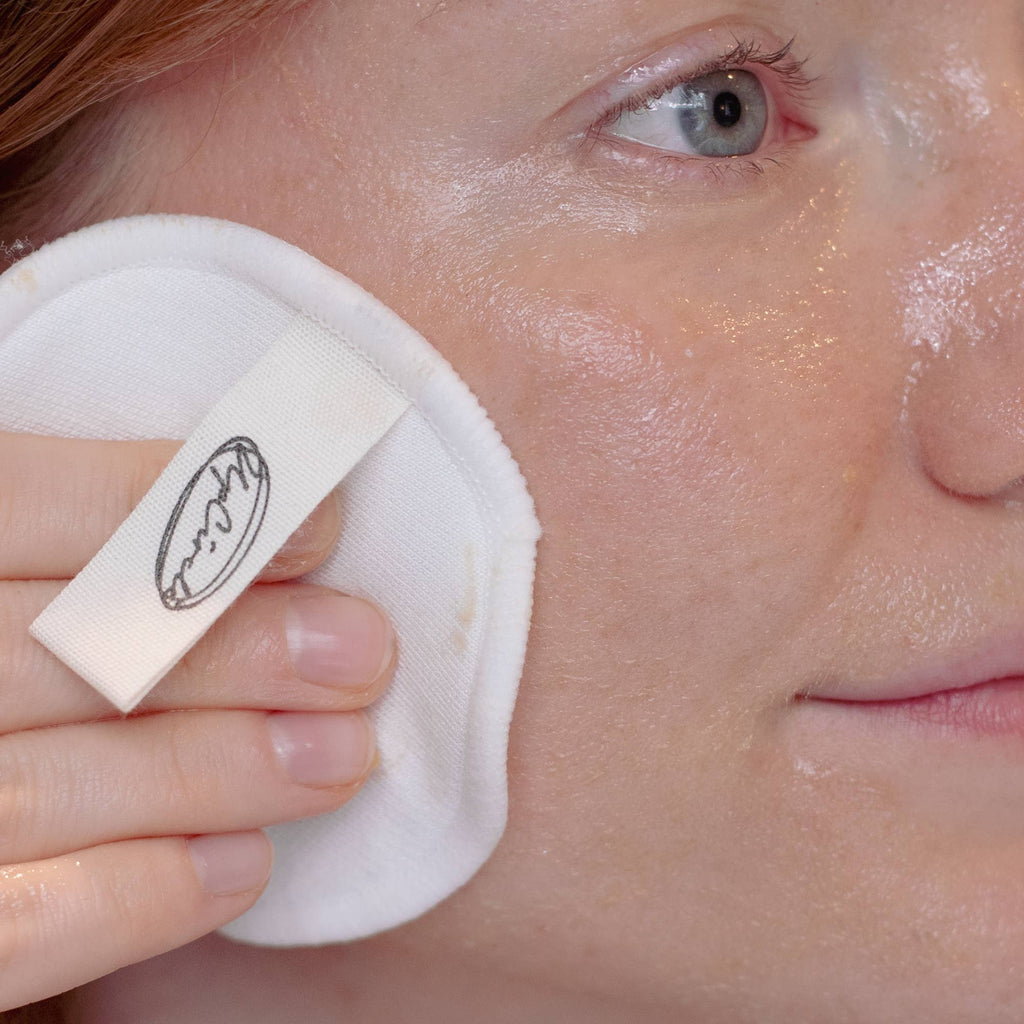 Sieben weiße, runde Make-up-Pads auf einem hellen Fliesenboden neben einem geöffneten Netzbeutel.
