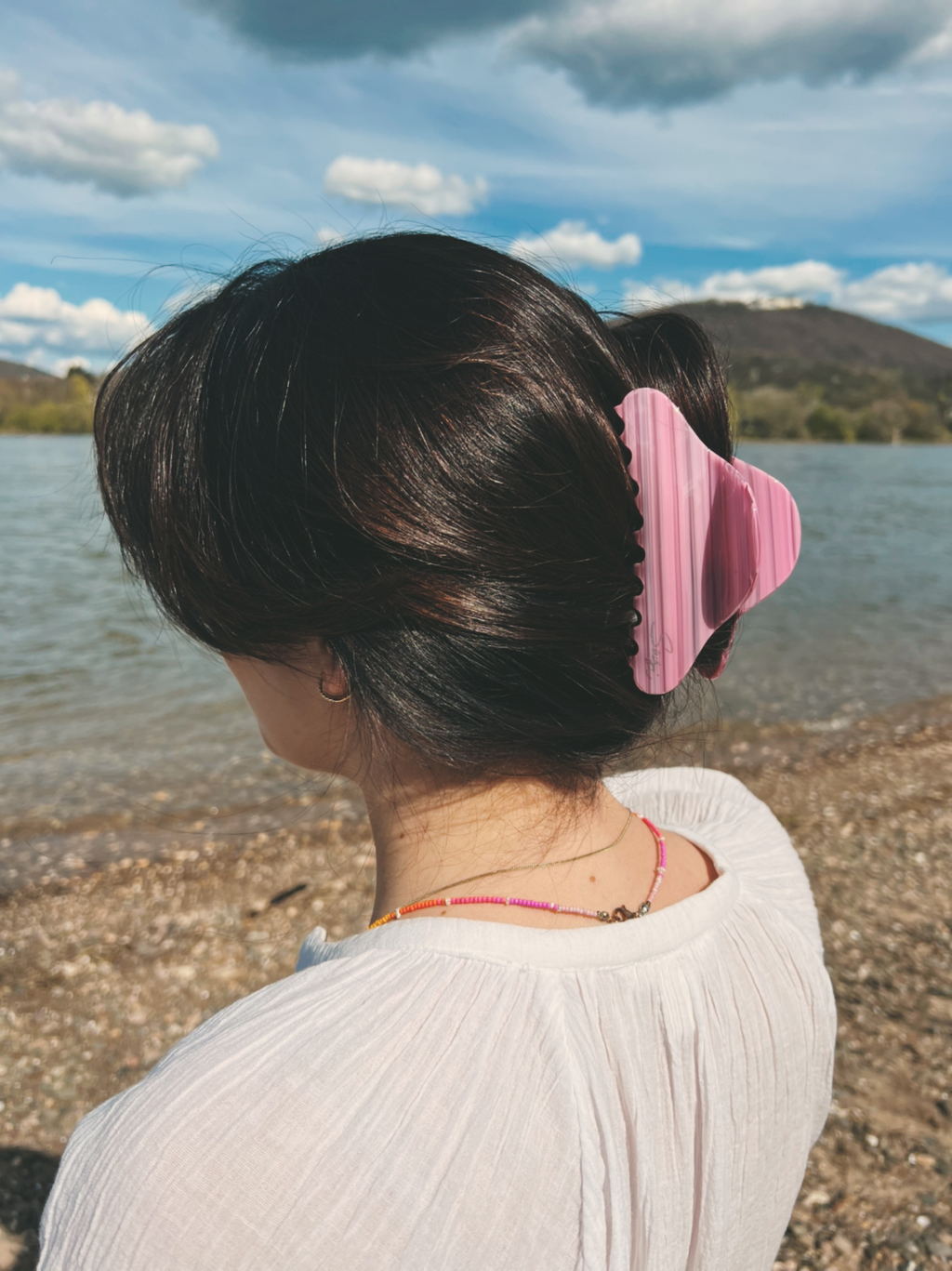 Rückansicht einer Frau mit dunklem Haar vor einer Wasserlandschaft, deren Frisur von der großen, pastellrosa gestreiften Cloud Haarklammer gehalten wird.